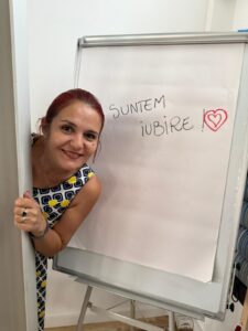 Bloggerița Elvira zâmbind larg, ascunsă parțial în spatele ei fiind un flipchart pe care scrie „SUNTEM IUBIRE!”, ilustrând entuziasmul pentru Spring SuperBlog 2026.